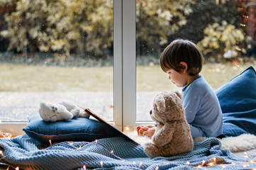 Cute little boy reading book with his teddy bear toy sitting cozy on pillows and knitted blanket near wet window with autumn garden at background. Cozy home. Winter holidays lifestyle.