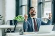 © LIGHTFIELD STUDIOS - happy businessman looking up and showing winner gesture while sitting at workplace
