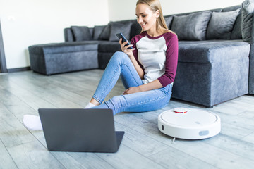 Young pretty woman sitting on the floor with laptop and control robot vacuum cleaner from the phone