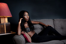 African American Woman Relaxing In Lingerie On Gray Sofa. Red Lamp On Table And Dark Background.  She Has Long Black Hair And Sensual Look. 