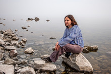 European Girl Tourist Sitting On A Large Stone Near The Water With Fog. Girl In A Striped Shirt And Pants. The Water Is Clear, Under Water Overgrown With Silt Stones.