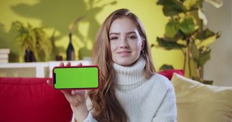Appealing cute ginger girl showing smartphone horizontal green screen mock-up smiling enjoying modern online shopping app using technology staying at home.