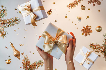 Wall Mural - Female hands holding gold gift box on light golden background with flying Christmas decorations, fir branches, balls, sparkles and confetti. Xmas and New Year holiday, bokeh, light. Selective focus