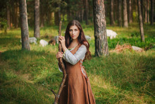 Young Beautiful Girl In Medieval Cowboy Clothes, With A Stick In Hand. Barefoot On The Ground. Against The Background Of The Forest And Green Grass. A Model With Clean Skin.