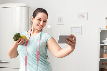 Canvas Print - Body Care. Chubby girl standing with tape measure around neck in kitchen taking selfie on smartphone posing to camera with broccoli and apple joyful