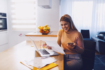 cute caucasian smiling blonde woman in sweater holding bills in one hand and in other smart phone. o