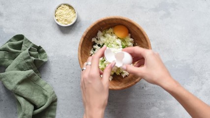 Wall Mural - Cooking process. Preparing zucchini fritters dough in a bowl. Woman chef add chicken egg into mixture of grated zucchini, herbs and goat cheese