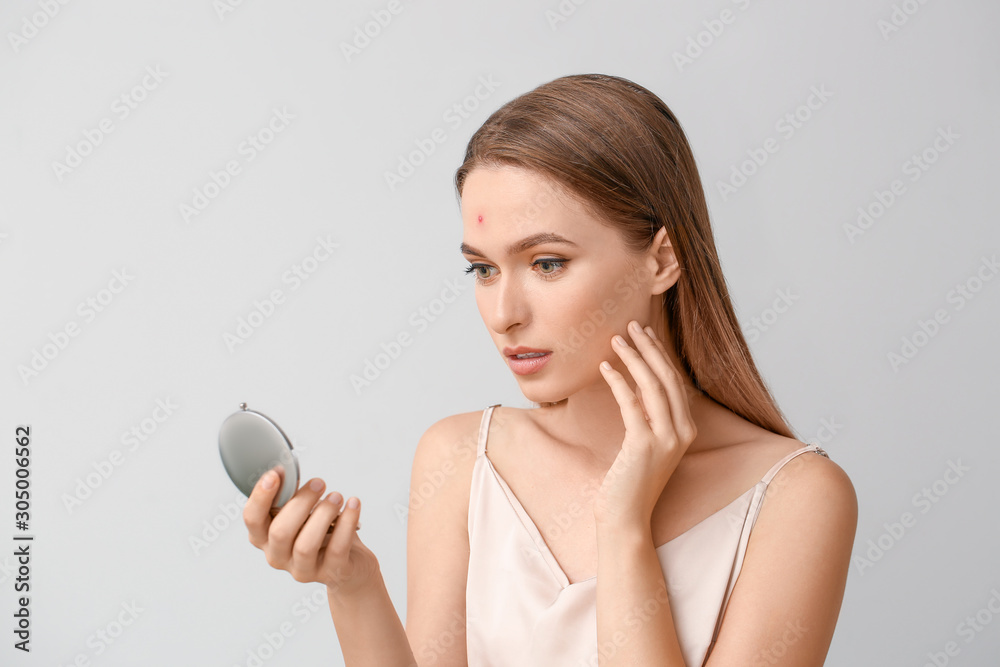 Portrait of young woman with acne problem looking in mirror on light background