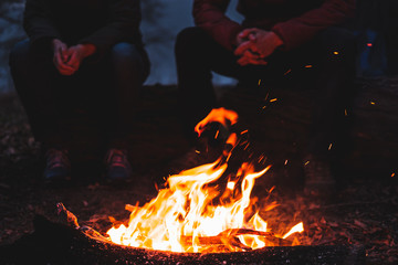 Two people sit by the bright bonfire at dusk. Spending nice time outdoors in chilly weather at a camping place - tranquil and peaceful scene