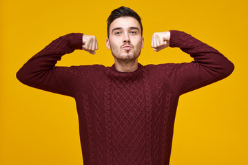 Wall Mural - Stylish attractive young bearded guy in knitted cozy sweater demonstrating strength, raising hands, keeping fists clenched, feeling confident and proud of himself. Confidence and power concept