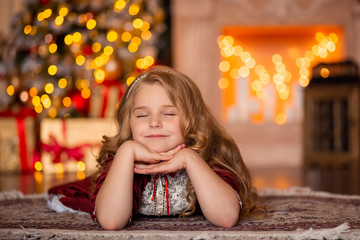 Wall Mural - A little beautiful girl with white hair and in a red dress lies on the carpet in the room. And dreams of gifts. behind her is a Christmas tree in lights and a fireplace.