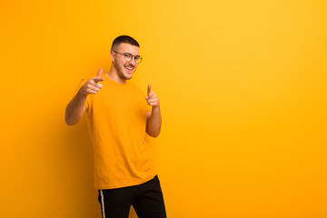 young handsome man smiling with a positive, successful, happy attitude pointing to the camera, making gun sign with hands against flat background