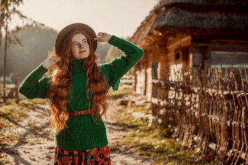 Outdoor autumn portrait of young happy redhead model, girl with natural freckled skin, long curly hair wearing orange hat, green knitted sweater, posing in beautiful old countryside. Copy, empty space