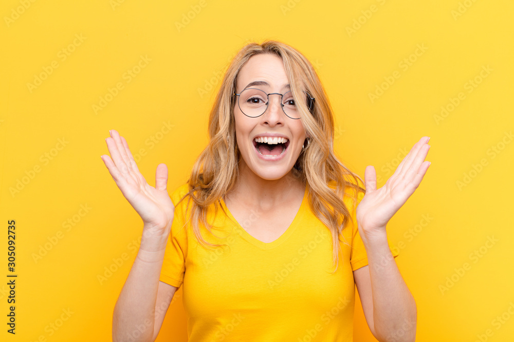 young pretty blonde woman looking happy and excited, shocked with an unexpected surprise with both hands open next to face against flat color wall - obrazy, fototapety, plakaty 