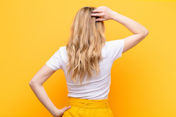 young pretty blonde woman feeling clueless and confused, thinking a solution, with hand on hip and other on head, rear view against flat color wall