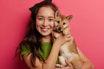 portrait of caring mixed race woman keeps pedigree puppy closely to face, embraces shiba inu dog, ha