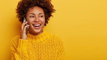 Wall Mural - Horizontal shot of optimistic curly woman has pleasant telephone conversation, wears knitted sweater, uses cellular, looks aside and smiles broadly, isolated over yellow wall, giggles at funny joke