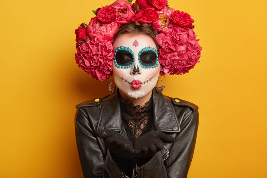 Photo of lovely woman blows mwah, keeps lips folded, wears creative makeup, prepares for carnival, prepares for Day of Dead, expresses love, poses against yellow background. Mexican female sends kiss