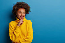 Positive Curly Young Woman Dressed In Yellow Comfortable Sweater, Holds Chin, Looks Aside With Dreamy Expression, Has Interesting Idea In Mind, Isolated Over Blue Background. Ethnicity Concept