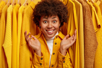 Happy Afro woman with toothy smile, raises palms and shows big variety of clothes in store, looks through yellow attires on hangers, prepares for going out, buys outfit on sale during Black Friday