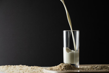 Pouring oat milk into glass on black background and oat flakes on table. Oat milk lactose free milk substitute concept. Copy space, soft shadows.