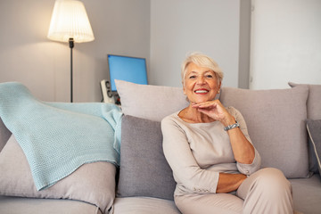 Indoor portrait of attractive happy 60 year old senior woman with pleasant smile relaxing on grey sofa in her modern apartment looking in anticipation while waiting for her children or grandchildren