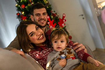 Wall Mural - Joyful family enjoying on Christmas Eve at home. Happy young parents with their baby boy enjoy Christmas time while relaxing on the sofa. Family prepare for Christmas