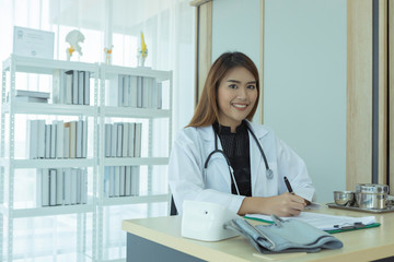 A beautiful young Asian doctor is writing a report on a patient's desk in the hospital with a smiling face.