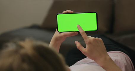Young woman lying on a couch and using smartphone with horizontal green screen