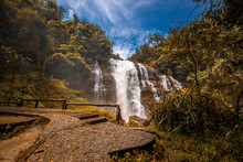 The Blurred Background Of Water Droplets From The Waterfalls That Cover The Area Around The Walkway, Surrounded By Large Trees And Fresh Cool Air While Visiting The Scenery.