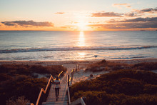 Young Woman Walking Down The Beach Staircase And Path To The Sea With The Orange Sunset Over The Beautiful Ocean In Perth, Western Australia.  