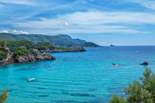 Landscape With Turquoise Calm Sea Water, Mountain With Rocky Hillside Covered With Green Trees And Bushes And Caves, Cruise Touristic Boats And Clouds On The Sky. Corfu Island, Greece. 