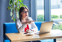 Portrait Of Shocked Beautiful Stylish Brunette Young Woman In Glasses Sitting, Pointing And Looking At Her Laptop Screen, Reading Unbelievable News. Indoor Studio Shot, Cafe, Office Background.