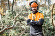 Waist-up portrait of a professional lumberman in harhat and protective workwear standing in the pine forest