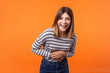 © khosrork - Portrait of overjoyed excited adorable young woman with brown hair in long sleeve striped shirt standing, holding her belly and laughing out loud. indoor studio shot isolated on orange background