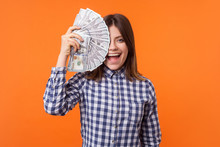 Half Face Portrait Of Amazed Attractive Brunette Woman Wearing Checkered Shirt Holding Dollars And Looking At Camera With Open Mouth, Lottery Winner. Indoor Studio Shot Isolated On Orange Background