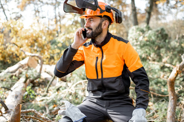 Canvas Print - Professional lumberman in protective workwear talking on phone, while sitting on the felled tree, resting after the hard work in the forest