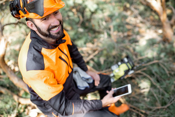 portrait of a professional lumberman in protective workwear sitting with mobile phone on the felled 