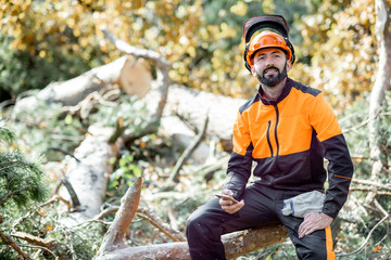 Wall Mural - Portrait of a professional lumberman in protective workwear sitting on the felled tree while resting after the hard work in the forest