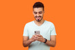 © khosrork - Portrait of cheerful brunette man with beard in white t-shirt using cellphone and smiling, reading good news message, enjoying mobile application. indoor studio shot isolated on orange background