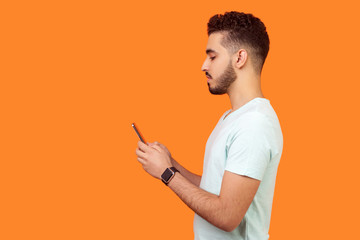 side view of serious calm brunette man with beard in white t-shirt using cellphone, texting chatting