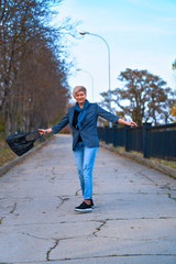 Wall Mural - Happy blond woman walking and spinning through autumn city park, dressed in blue jeans and jacket