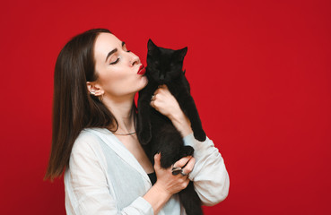 Beautiful woman in white shirt hugs and kisses black cat on red background. A woman loves her pet. Portrait of a girl with a cat in her arms. Isolated on red.