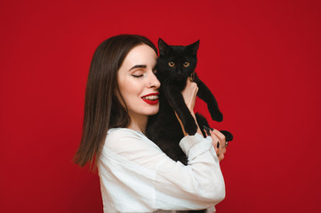 Smiling woman in light casual clothes stands on a red background with a black cat in her hands, hugging the pet with his eyes closed.Happy black cat and owner isolated on red background. Pets concept