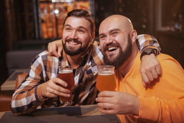 Happy bearded man enjoying drinking beer with his best friend. Male friends hugging, holding up their beer glasses, celebrating at the bar