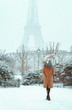 A young woman in a beige coat walks under an umbrella in a snowy winter Paris against the backdrop of the Eiffel Tower. Romantic brunette girl is walking in the background of falling snow in Paris. 