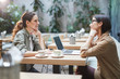 © Seventyfour - Side view portrait of two modern young women sitting at table in cafe while discussing business during meeting on outdoor patio, copy space