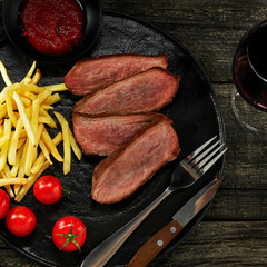 Wall Mural - Delicious medium rare steak with French fries and fresh cherry tomatoes on a black plate. Classic meat meal with a glass of red wine. Top view, directly above shot.