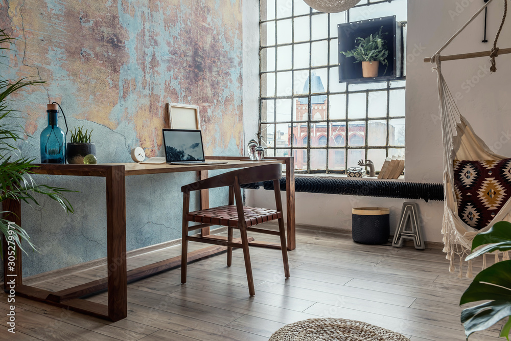 Stylish interior design of office space in loft apartment with wooden desk, chair, office supplies, laptop, plants, hammock  and elegant accessories. Modern home office decor. Bright space. Template. - obrazy, fototapety, plakaty 