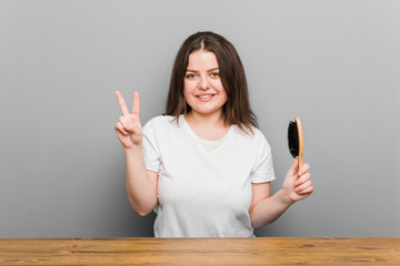 Wall Mural - Young plus size curvy woman holding an hairbrush showing number two with fingers.